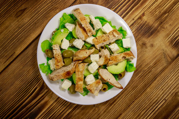 Tasty salad with fried chicken breast, green olives, feta cheese, avocado, lettuce and olive oil on wooden table. Top view