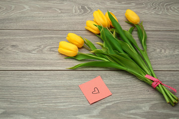 Yellow tulips lie on a gray wooden table next to a sticker on which a heart is drawn.