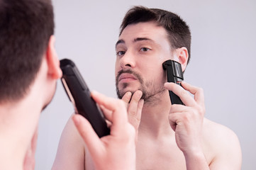 A young guy shaves in the bathroom. The guy shaves his beard with a trimer. Care beard.