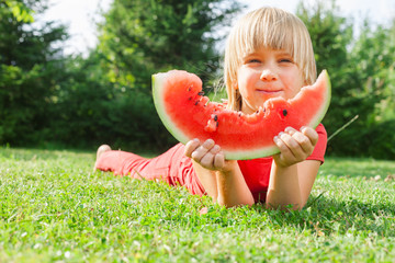 Happy little girl eating slice of melon lying in a summer garden