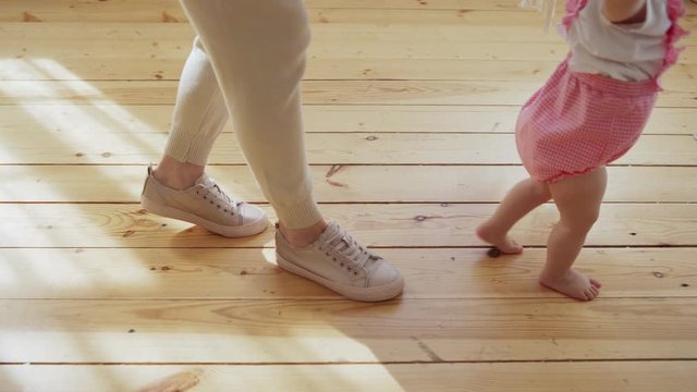 Tracking shot of unrecognizable mother helping her lovely baby girl to make her first steps on hardwood floor