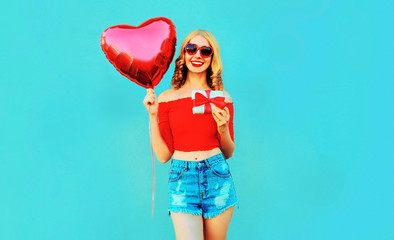 portrait happy smiling young woman holding gift box, red heart shaped air balloon on colorful blue background