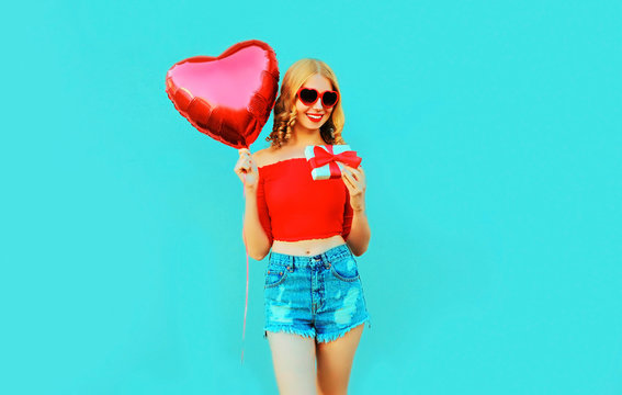 portrait happy smiling young woman holding gift box, red heart shaped air balloon on colorful blue background