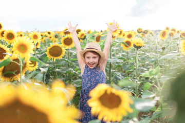 Funny kid in sunflowers in summer vacation. Child girl in straw hat and blue dress is smiling, laughing. Concept of holiday, happiness and enjoying life. Lifestyle, authentic moments, emotions.