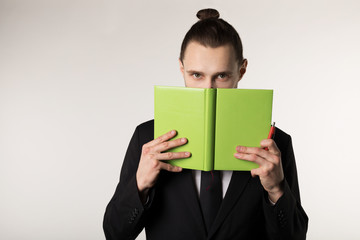 Young man in black suit with trendy hairstyle hides his face behind the green book