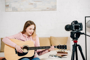 Teenage blogger sitting on bed and playing acoustic guitar in front of camera