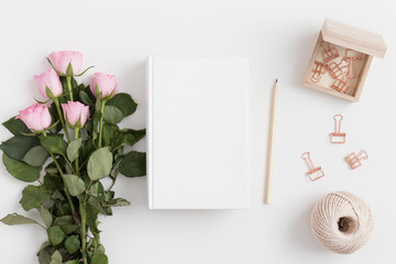 White book mockup with a bouquet of pink roses and workspace accessories on a white table.