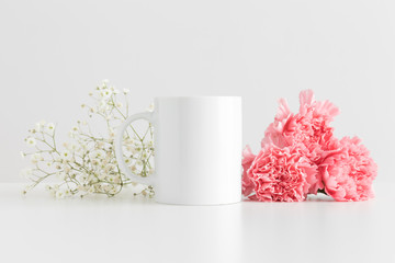 Mug mockup with a bouquet of pink carnations and a gypsophila on a white table.