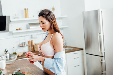 Sensual woman in lingerie and shirt holding cherry tomatoes in kitchen