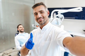 Handsome happy young man doctor in medical dentist center pointing take a selfie by camera.