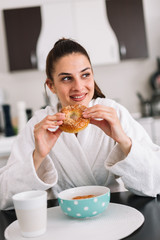 Woman having a breakfast in a robe at home. Girl eating a bagel at home.
