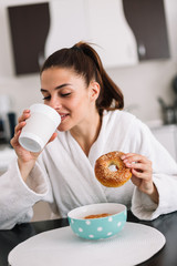 Woman having a breakfast in a robe at home. Girl eating a bagel at home.