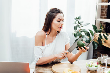 Sexy girl cutting avocado with knife at wooden table in kitchen