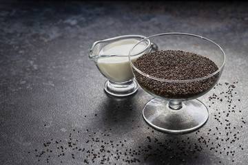 Chia seeds in a glass vase and coconut milk against a dark stone surface with scattered grains.