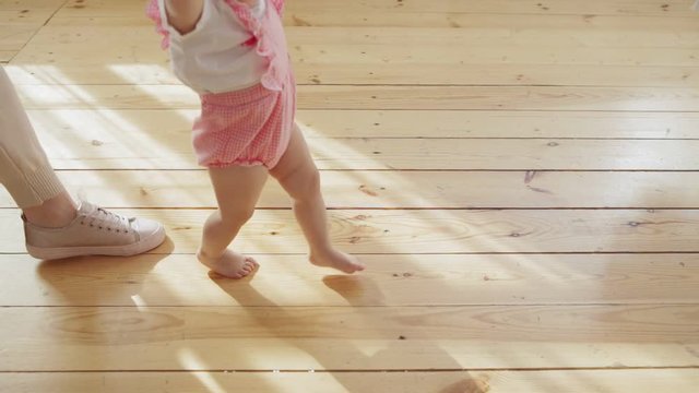 Unrecognizable caring mother helping her toddler daughter to make her first steps on wooden floor in slow motion