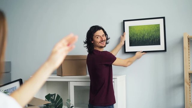Young couple man and woman is choosing place for picture on wall in new house after relocation, girl is making frame with fingers then showing thumbs-up.
