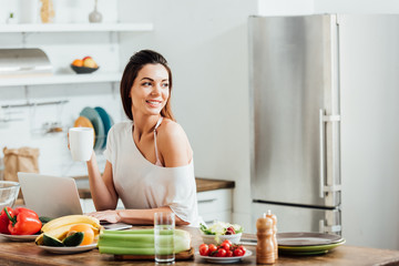 Charming girl drinking coffee while using laptop in kitchen