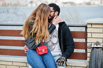 Cool multiracial couple sitting against lake background and hugs together.