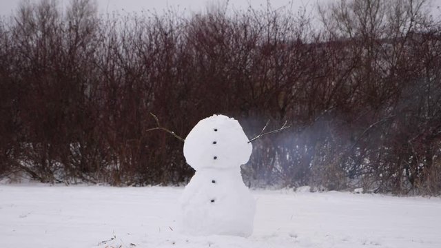 Snowman just loose his head, light smoke fly beside rest of body, two large snowballs stay against dark naked bush twigs. Slow motion shot, fume cloud and dissolve with time