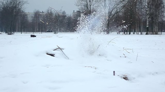 Small explosion inside snowpile, cracker cylinder and debris fly away, close slow motion shot. Winter park area, trees and buildings seen on background. Petard blow up inside heap of wooden chips