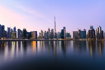 Fototapeta na wymiar Panoramic view of the illuminated Dubai skyline during sunset with the magnificent Burj Khalifa and many others skyscrapers reflected on a silky smooth water flowing in the foreground. Dubai.