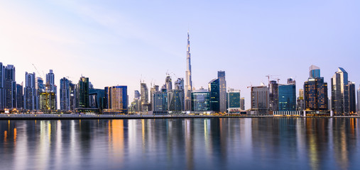 Obraz premium Panoramiczny widok na rozświetloną panoramę Dubaju podczas zachodu słońca ze wspaniałym Burdż Chalifa i wieloma innymi drapaczami chmur odbijającymi się w jedwabiście gładkiej wodzie płynącej na pierwszym planie. Dubai.