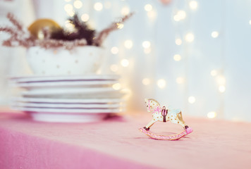 Composition with vintage Christmas tree toy porcelain figurine of a rocking horse and dishes on the pink table on the background of blurred lights. Festive layout. Soft selective focus. Copy space.