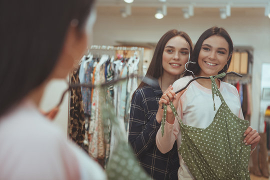Cheerful beautiful woman smiling, helping her best friend choosing a new dress to buy. Two female friends examining a dress in front of the mirror at clothing store. Gorgeous woman shopping with frien