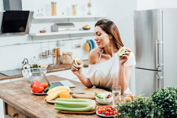 Pretty stylish girl holding cut avocado near table in kitchen