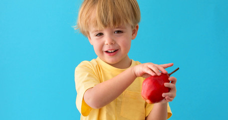 A sweet little boy smiles whith red pear. Kid shows the pear to camera on blue background