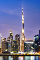 Atemberaubende Aussicht auf die beleuchtete Skyline von Dubai bei Sonnenuntergang mit dem prächtigen Burj Khalifa und vielen anderen Gebäuden und Wolkenkratzern, die sich auf einem seidig glatten Wasser im Vordergrund widerspiegeln.