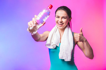 Resting time. Sporty girl with bottle of water and towel on her shoulders. Photo of fitness model isolated on white background. Sport and healthy lifestyle. Thumbs up.