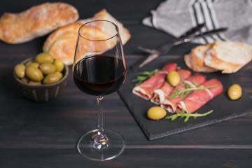 Glass of red wine with slices of cured ham or Spanish jamon serrano or Italian prosciutto crudo, bread, green olives and arugula