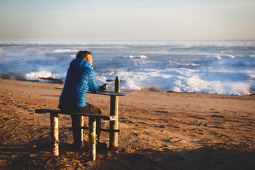 A man sits on a bench on a frozen shore. Thermos on the table. Sunny evening. The background is blurred
