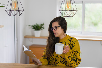 Mulher de óculos segurando caneca branca numa mão enquanto segura um caderno aberto na outra, numa bancada de madeira. 