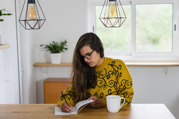 Mulher de óculos a escrever no seu caderno com uma caneca branca ao lado em cima da bancada de madeira.