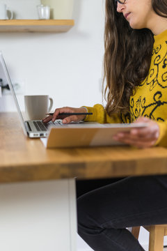 Mulher a trabalhar no seu computador port&aacute;til vista de lado. A segurar um caderno de apontamentos e com uma caneca branca do seu lado direito.