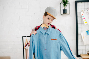 Teenager kid in cap holding hanger with blue shirt at home