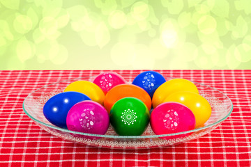 Easter decorations background. Colorful easter eggs in a glass bowl on a red checkered tablecloth against abstract light green background. Religion and culture concept.