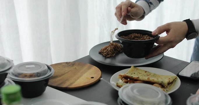 Man in kitchen area puts buckwheat porridge from black bowl on plate