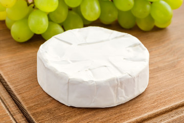 Close-up of whole head of camembert and sweet green grapes on a brown wooden cutting board. Soft...