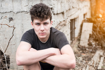 A teenager with a sad serious face sits near the old house and regrets having quarreled with his friends