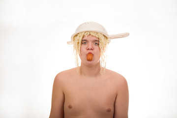A young, fat, cheerful, smiling boy eating sausage and having a colander and spaghetti on his head. Plump body without clothing. White background. Portrait photo.