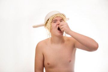 A young, fat, cheerful, smiling boy eating pasta and having a colander and spaghetti on his head. Plump body without clothing. White background. Portrait photo.