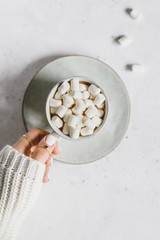 Girl's hand holds a cup of hot chocolate with marshmallow over textured white background. Minimalist style, top view, copy space.