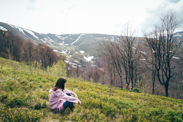Fototapeta na wymiar Tourist girl in pink rain jacket sitting on the grass looking at mountains surrounded by forest, enjoying silence and harmony of nature