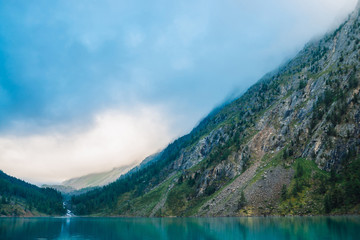 Naklejka na ściany i meble Giant cloud above mountainside with trees in sunlight. Amazing mountain lake. Mountain range under blue cloudy dawn sky. Wonderful rocks. Morning landscape of highland nature. Low cloud in sunny light