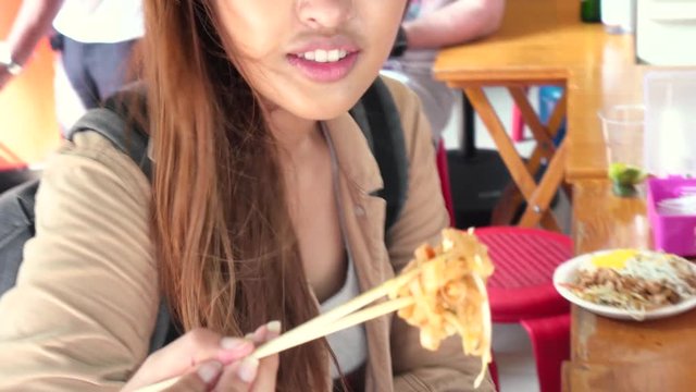 Young Asian tourist woman eating pad thai noodle, traditional street food in Thailand and looking at camera