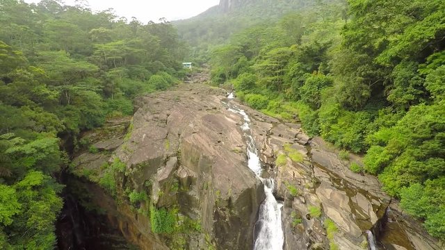 Backwards drone shot of the Bambarakanda Falls, Sri Lanka, surrounded by bright green trees, on a cloudy and foggy day
