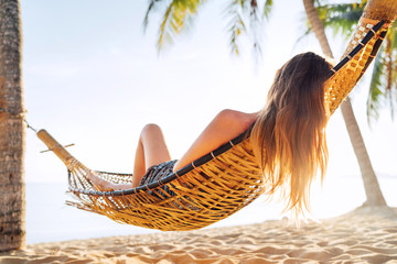 Blonde longhaired woman relaxing in hammock hinged between palm trees on the sand beach